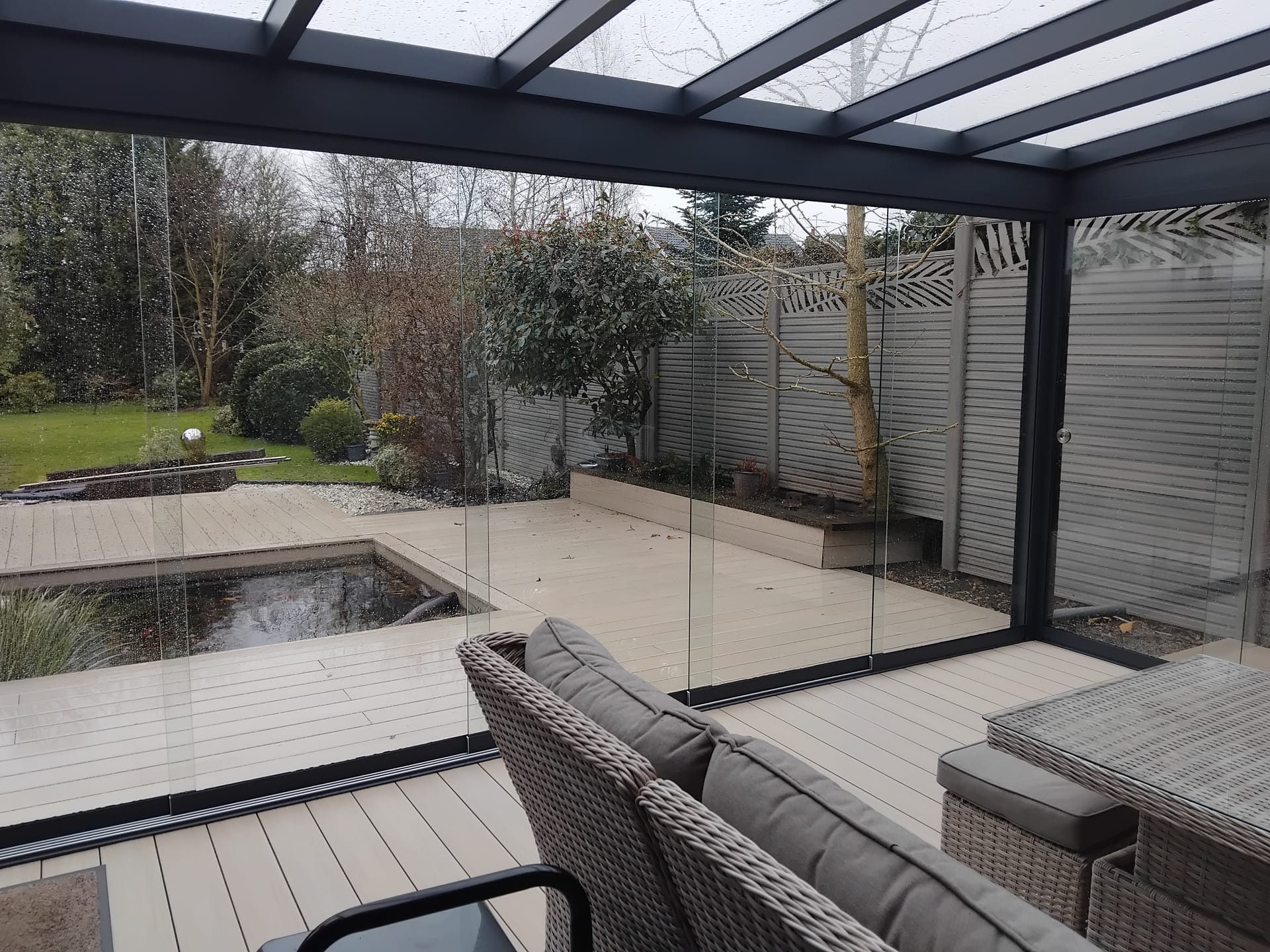 Inside view from a glass garden room with frameless sliding glass walls, looking out over decking, a pond and garden